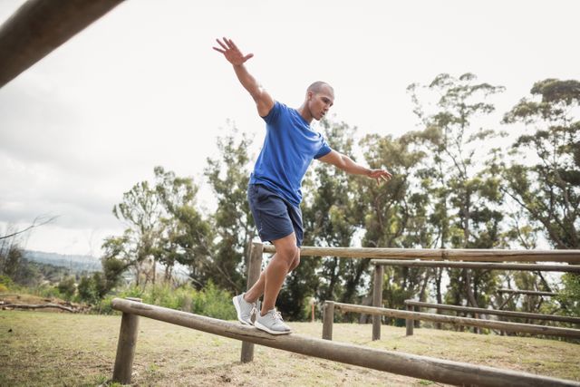This image shows a fit man balancing on hurdles during an obstacle course training at a boot camp. The setting is outdoors, surrounded by trees and nature, emphasizing physical fitness and determination. This image can be used for promoting fitness programs, boot camps, outdoor activities, and healthy lifestyles.