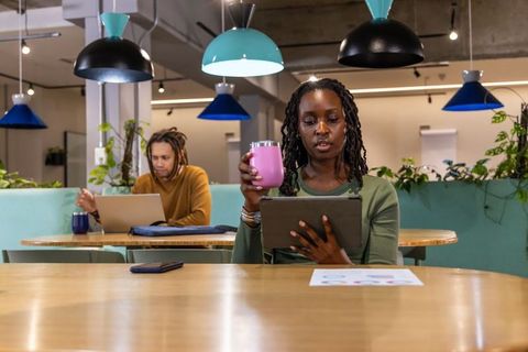 African American woman working in coworking space holding tablet and pink tumbler