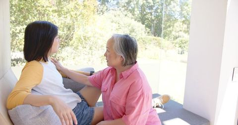 Asian mother comforting teenage daughter on sunny window seat, close family conversation