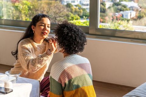 Friends applying makeup by sunlit window in cozy knitwear, sharing beauty routine