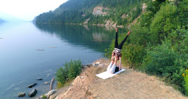 Photo shows a couple practicing acro yoga on a mat by a scenic lake, surrounded by lush green trees and mountains. The calm waters and natural beauty suggest tranquility and relaxation. This image can be used for promoting outdoor fitness, yoga retreats, wellness blogs, healthy lifestyle products, or for travel and adventure advertisements.