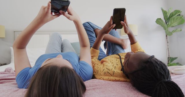 Two teenage friends comfortably lying on a bed and using smartphones in a cozy bedroom. Great for illustrating concepts of friendship, modern technology, social media, and communication. Suitable for use in advertisements, social media content, promotion of mobile apps, or articles on teenage life and technology usage.