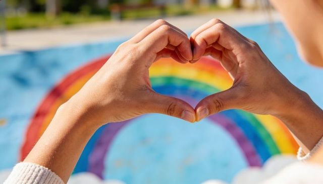 Hands forming heart framing vibrant rainbow mural in sunlit pool closeup with knit sweater cuffs