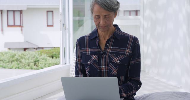 Mature woman using laptop indoors near window, dressed casually in plaid shirt. Bright sunlight filtering through. Ideal for concepts of technology use among elders, remote work, relaxation, sunny day indoors.