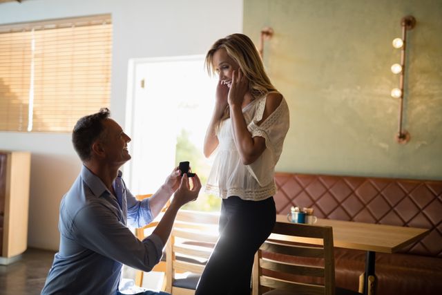 Man kneeling and presenting engagement ring to surprised woman in restaurant. Ideal for use in articles or advertisements about proposals, engagements, romantic moments, and relationship milestones.