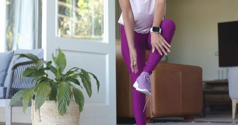 Woman stretching and balancing on one leg in bright living room wearing purple leggings and smartwat