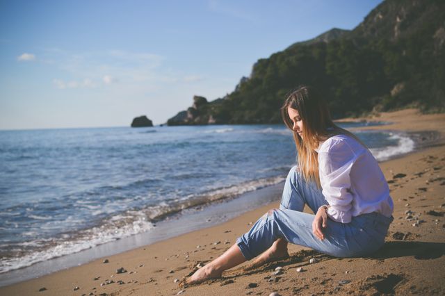 Young woman with long hair sitting on a sandy beach, enjoying a quiet moment with serene nature around. Waves gently hitting the shore, clear blue sky, and lush greenery in background. Perfect for themes related to relaxation, contemplation, travel, nature exploration, and peaceful moments.