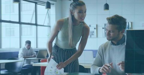 Young woman presenting printed charts to colleague while pointing at monitor in coworking space