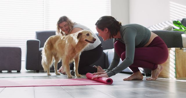 Two women are practicing yoga with their dog in a bright living room. One woman rolls a yoga mat while the other smiles at the dog. This scene is perfect for illustrating a home workout, healthy living, and the inclusion of pets in daily activities. It is a great fit for fitness blogs, home exercise guides, pet-friendly workout routines, and lifestyle magazines.