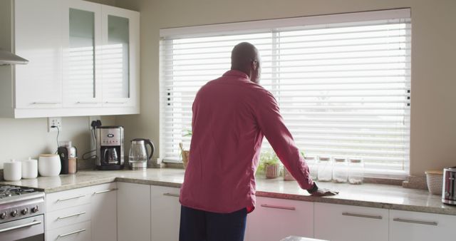 Man standing in a contemporary kitchen looking outside the window with blinds partially open. Kitchen features coffee machine, countertop, cabinets, and a bright atmosphere. Ideal for concepts related to everyday home life, morning routines, contemplation, and modern living spaces.