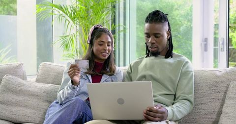 Diverse couple shopping online with laptop and credit card on bright modern home sofa