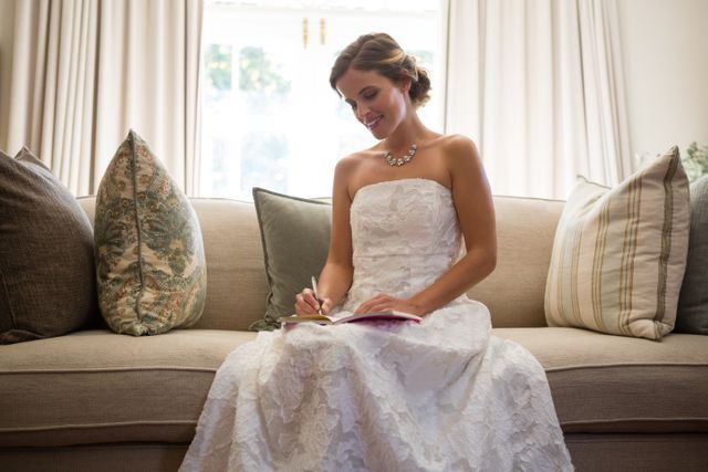 Bride writing in diary while sitting on sofa at home, capturing a personal and intimate moment on her wedding day. Ideal for use in wedding planning materials, bridal magazines, lifestyle blogs, and advertisements for wedding-related products and services.