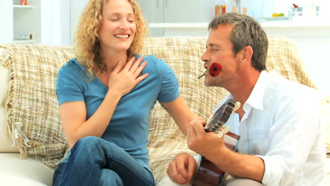 Man Serenading with Guitar Sitting Next to Smiling Woman at Home
