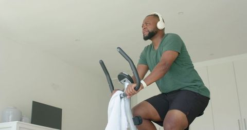 African american man cycling on stationary bike at home wearing headphones and smartwatch