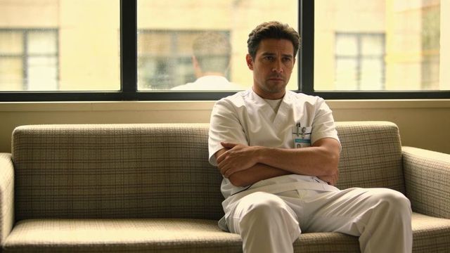 Contemplative male doctor in white scrubs sitting on clinic sofa by window with id badge