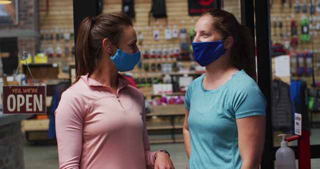 Caucasian female shopkeepers in face masks in the doorway of sports shop with arms crossed. fitness and leisure retail during coronavirus covid 19 pandemic.