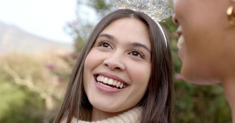 Smiling diverse women wearing silver tiara and cream scarf celebrating friendship outdoors
