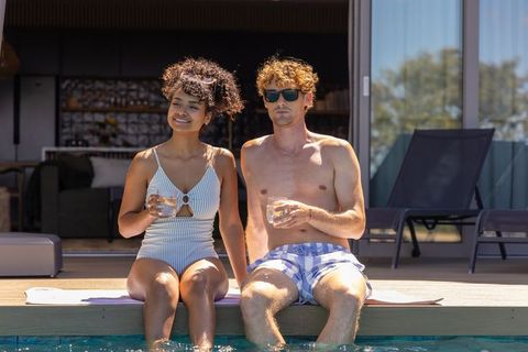 Young couple relaxing poolside, dipping legs in water while holding drinks on sunny deck