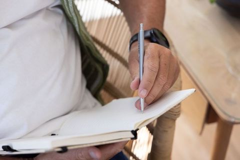 Mature man writing in lined notebook on woven chair at home office with smartwatch
