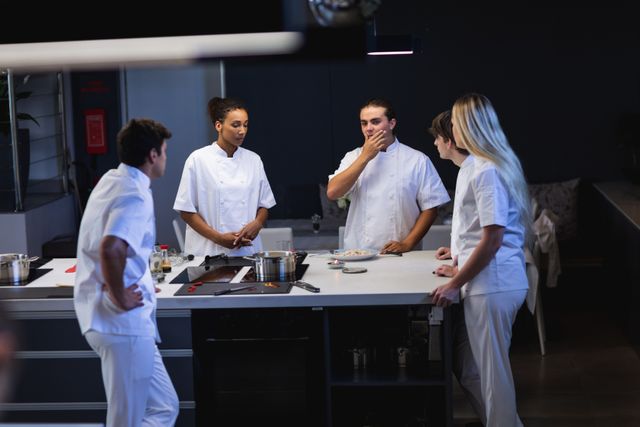 Multi ethnic group of chefs cooking in a modern busy kitchen, standing round table talking. Cookery class at a restaurant kitchen. Workshop cooking food.