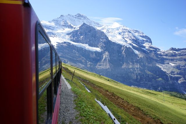 Red train traveling through scenic alpine region with snow-capped mountains and clear blue sky. Ideal for concepts of adventure, nature, travel, and tourism.