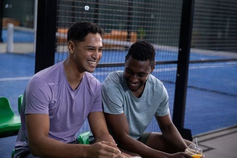 Laughing teammates sitting on bench at indoor court holding drinks, sports camaraderie