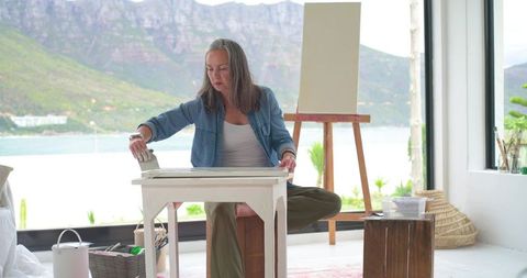 Senior woman painting at table by lake, coastal studio with easel and mountain view