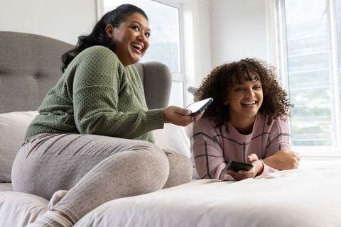 Diverse mom and teen daughter laughing together on bed, sharing phones, cozy loungewear
