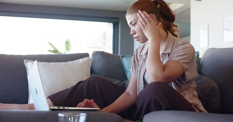 Young woman sitting on sofa holding head while typing on laptop in modern living room
