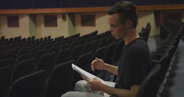 Young man sitting alone in auditorium reading script. Ideal for concepts of theater, rehearsal, practicing lines, solitude, focus, preparation for performance, or student practicing speech.
