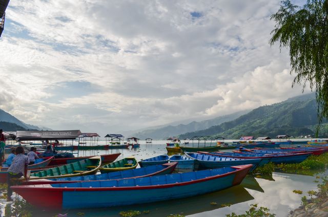 Lakeside filled with colorful rowboats under partly cloudy sky with mountains in background. Perfect for travel blogs, nature magazines, and peaceful retreat advertisements, highlighting serene outdoor experiences and scenic lakefront views.