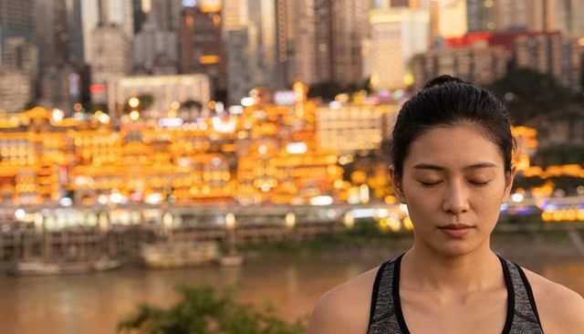 Woman practicing mindful breathing riverside at dusk with vibrant lit city skyline