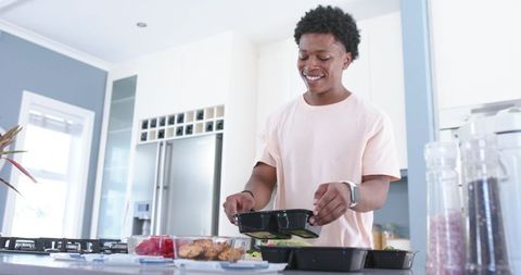 Young man packing healthy meal prep containers on kitchen island with berries and chicken