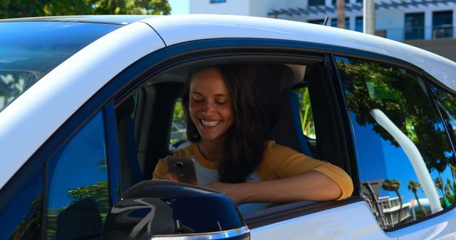 A woman is smiling while using her smartphone inside a car on a sunny day. The casual and relaxed scene depicts a modern lifestyle and connects well to themes of technology, communication, or outdoor activities. Ideal for advertisements relating to mobile technology, apps, or automotive services.