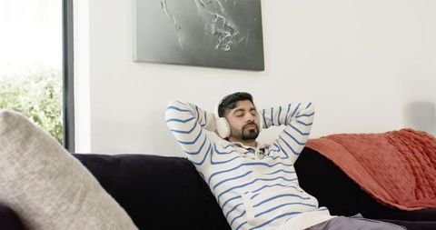 Indian man relaxing on sofa listening to music with headphones in bright modern living room