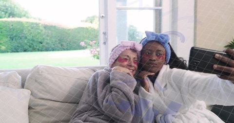 Two friends taking selfie during home spa night in cozy robes and eye patches