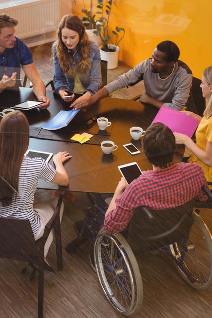 Group of diverse business professionals having a discussion around a table in a modern office setting. One team member is in a wheelchair, highlighting disability inclusion. Useful for depicting teamwork, professional environment, and inclusive workplaces in corporate settings.