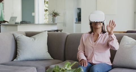 Mature woman experiencing virtual reality wearing white VR headset gesturing on gray sofa