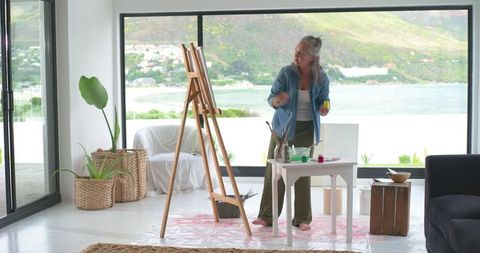 Mature woman painting barefoot on easel in bright seaside home studio, creative art lifestyle