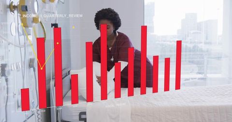 African american nurse straightening hospital bed while reviewing financial chart overlay