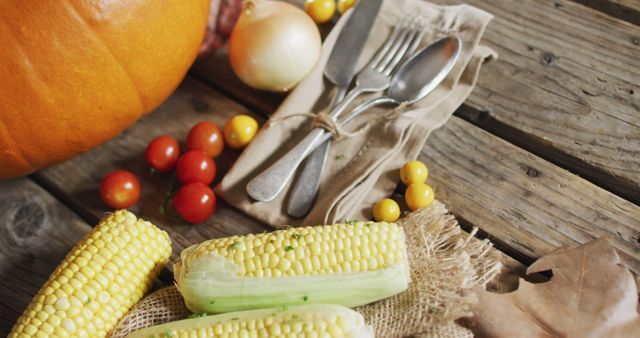 This image features a rustic autumn table arranged with fresh produce including corn, cherry tomatoes, an onion, a pumpkin, and cutlery on a wooden surface. The mix of seasonal vegetables represents the bountiful harvest. This can be used for content related to fall celebrations, farm-to-table dining, seasonal recipes, organic markets, and food-related seasonal advertisements.