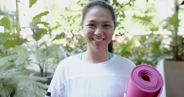 A smiling woman holding a pink yoga mat, standing in an lush outdoor setting. Perfect for use in fitness promotions, healthy lifestyle campaigns, yoga and wellness blogs, exercise or sports-related advertisements, and nature or relaxation themed content.