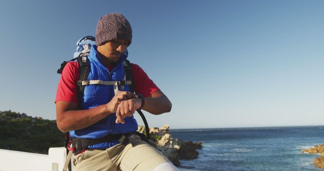 Image shows hiker checking smartwatch while taking a break by coastal trail on sunny day. He is wearing hiking gear and backpack. Useful for advertisements related to outdoor adventures, technology, fitness, and exploring nature.