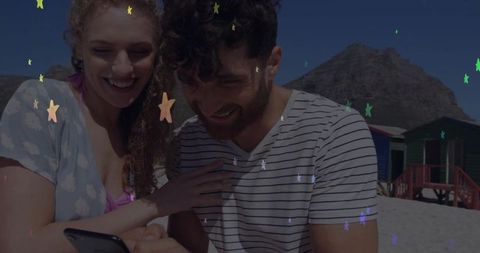 Smiling couple leaning together on beach looking at phone with colorful star overlay
