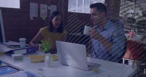 Discussing project at laptop while holding coffee cup, collaborating in modern loft office