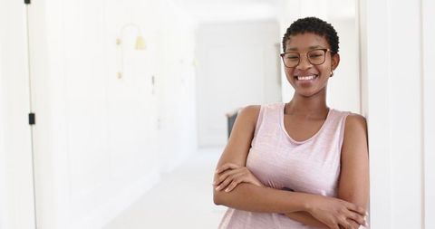 Smiling young woman leaning in bright minimalist hallway with arms crossed and glasses