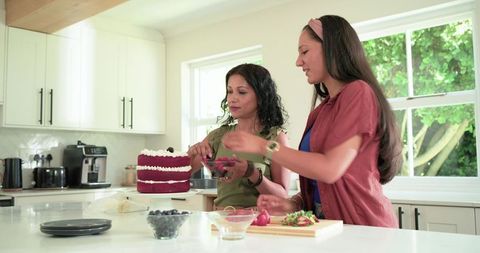 Mother daughter decorating red velvet layer cake together in sunlit modern kitchen