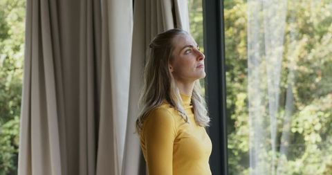 Contemplative woman gazing out window in mustard turtleneck natural light minimalist interior