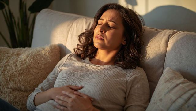 Woman holding abdomen while lying on sunlit couch in cozy living room, experiencing abdominal pain
