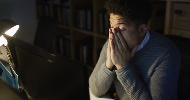 Young man appearing stressed and exhausted while working late at his office desk. Man rubbing his face with hands, technical looking stressed. Ideal for illustrating themes related to workplace stress, overwork, night shifts, and demanding corporate environments.